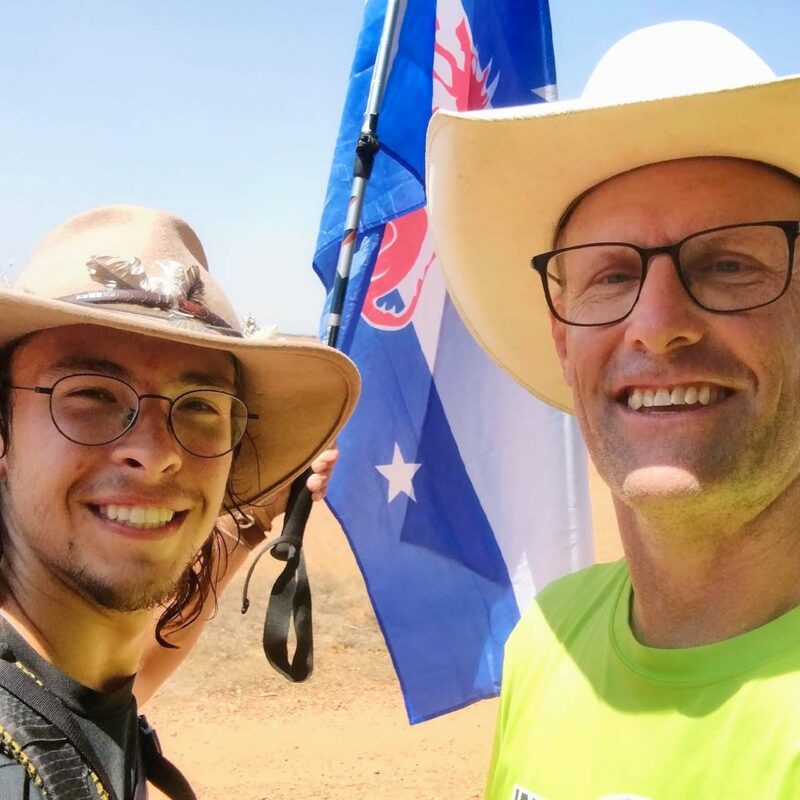 One older man and one younger man, both wearing cowboy-style hats, on a hiking trail carrying a Beta Theta Pi flag