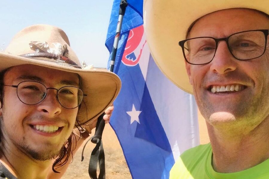 One older man and one younger man, both wearing cowboy-style hats, on a hiking trail carrying a Beta Theta Pi flag