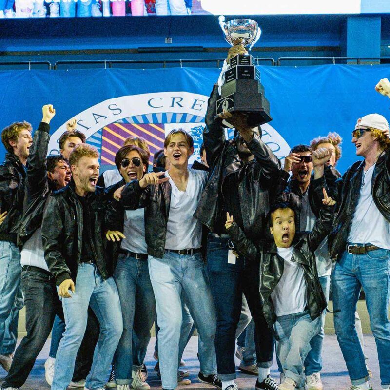A group of collegiate men in leather jackets hoist a large trophy overhead in celebration